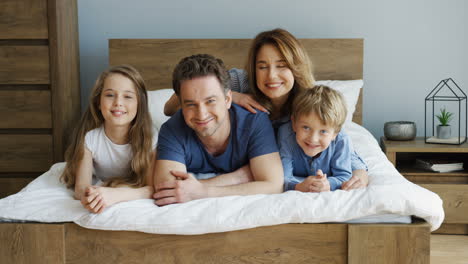 Cheerful-parents-and-their-sons-lying-on-the-bed-and-smiling-at-camera-in-the-morning