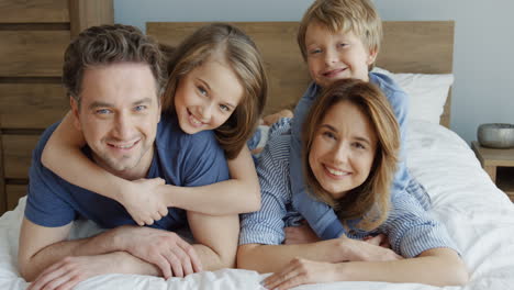Close-up-view-of-cheerful-parents-and-their-sons-lying-on-the-bed-and-smiling-at-camera-in-the-morning