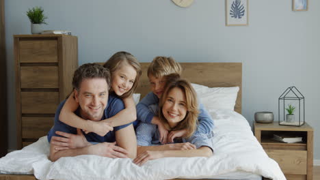 Fröhliche-Eltern-Und-Söhne-Liegen-Auf-Dem-Bett-Und-Lächeln-In-Die-Kamera