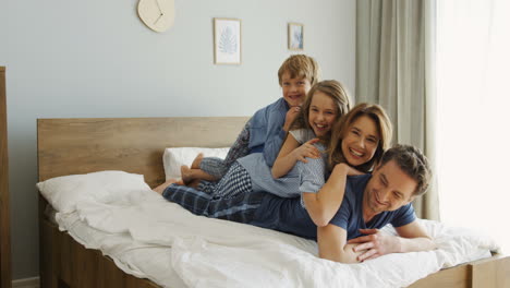 Cheerful-parents-and-sons-lying-on-the-back-of-each-other-on-the-bed-and-smiling-at-the-camera