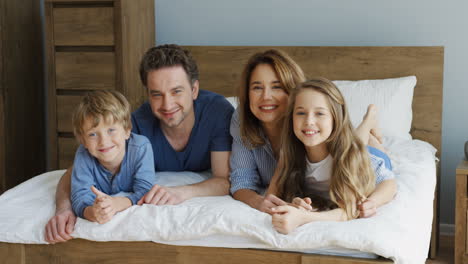 Close-up-view-of-cheerful-parents-and-their-sons-lying-on-the-bed-and-smiling-at-camera-in-the-morning
