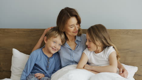 Young-beautiful-mother-sitting-on-the-bed-under-the-blanket-with-her-son-and-daughter,-hugging-and-kissing-them-in-the-morning