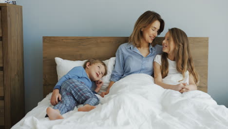 Young-beautiful-mother-sitting-on-the-bed-under-the-blanket-and-talking-with-her-son-and-daughter-in-the-morning