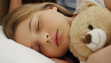 Close-up-view-of-cute-blonde-little-girl-sleeping-on-the-bed-while-hugging-her-teddy-bear-in-the-morning