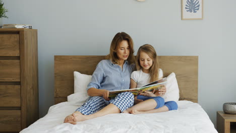 Young-beautiful-mother-lying-on-the-bed-under-the-blanket-and-reading-a-book-with-her-daughter-in-the-morning