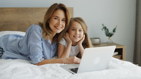 Young-beautiful-mother-lying-on-the-bed-with-her-daughter-in-front-of-laptop-while-smiling-at-camera-in-the-morning