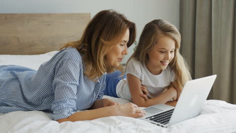 Young-beautiful-mother-lying-on-the-bed-with-her-daughter-and-watching-something-on-laptop-in-the-morning