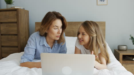 Close-up-view-of-happy-young-woman-lying-with-her-lovely-daughter-watching-something-on-laptop-lying-on-the-bed-in-the-morning