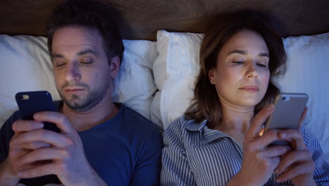 Top-view-of-caucasian-couple-lying-on-the-bed-and-using-smartphones-at-night