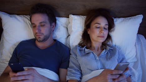 Top-view-of-caucasian-couple-lying-on-the-bed-and-using-smartphones-at-night
