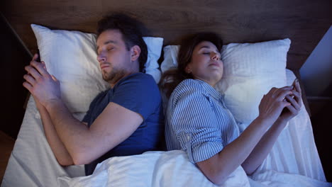 Top-view-of-caucasian-couple-lying-on-the-bed-back-to-back-and-using-smartphones-at-night,-then-they-turn-to-each-other-for-a-second