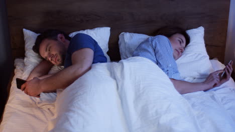 Top-view-of-caucasian-couple-lying-on-the-bed-back-to-back-and-using-smartphones-at-night
