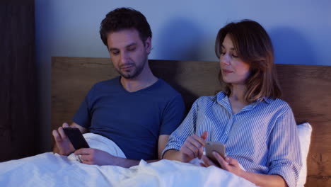 Kaukasisches-Paar,-Das-Auf-Dem-Bett-Sitzt-Und-Nachts-Smartphones-Benutzt