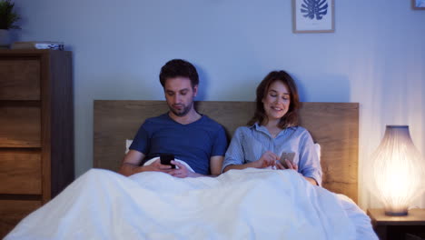 Kaukasisches-Paar,-Das-Auf-Dem-Bett-Unter-Den-Decken-Sitzt-Und-Nachts-Smartphones-Benutzt