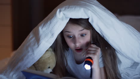 Close-up-view-of-cute-little-girl-using-a-flashlight-and-reading-a-book-with-surprised-expression-under-the-blanket-at-night