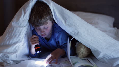 Nahaufnahme-Eines-Süßen-Kleinen-Jungen,-Der-Eine-Taschenlampe-Benutzt-Und-Nachts-Ein-Buch-Unter-Der-Decke-Liest