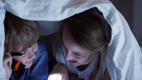 Close-up-view-of-cute-little-boy-and-her-sister-using-a-flashlight-talking-and-staring-at-something-under-the-blanket-at-night