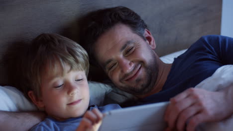 Top-view-of-happy-caucasian-father-lying-on-the-bed-with-his-cute-little-son-scrolling-and-taping-on-the-tablet-at-night