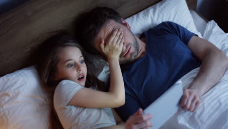 Top-view-of-little-girl-lying-on-the-bed-with-her-father-and-watching-video-on-the-tablet,-then-she-covers-the-father-eyes-because-he-is-scared