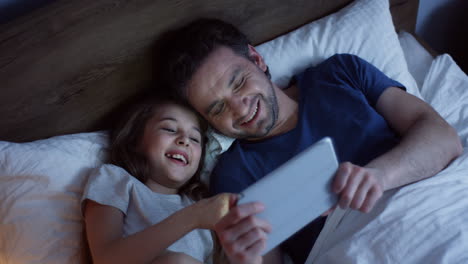 Top-view-of-little-girl-lying-on-the-bed-with-her-father-and-watching-video-on-the-tablet,-laughing-and-having-fun