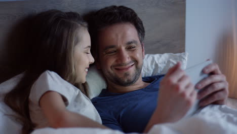 Little-girl-lying-on-the-bed-with-her-father-and-watching-video-on-the-tablet,-laughing-and-having-fun