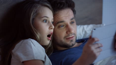 Close-up-view-of-little-cute-girl-lying-on-the-bed-before-sleeping-with-her-handsome-father-and-watching-scary-video-on-the-tablet