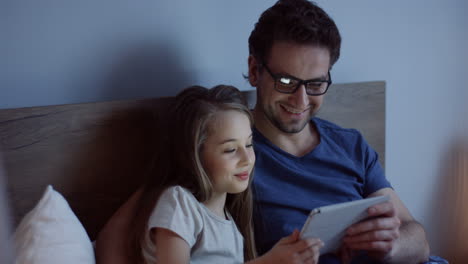 Handsome-Caucasian-man-in-glasses-with-his-little-pretty-blond-daughter-watching-something-on-the-tablet-and-smiling-while-they-sitting-on-the-bed-at-night