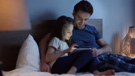 Caucasian-father-lying-on-the-bed-with-his-cute-little-daughter-scrolling-and-taping-on-the-tablet-at-night
