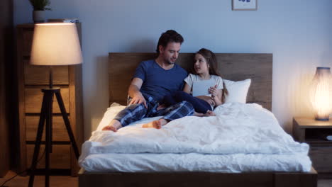 Caucasian-father-lying-on-the-bed-with-his-cute-little-daughter-scrolling-and-taping-on-the-tablet-at-night