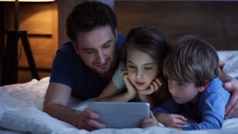 caucasian-father-and-his-little-son-and-daughter-lying-on-the-bed-and-watching-something-on-the-tablet