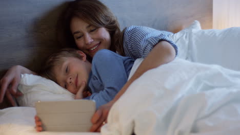 Little-cute-boy-on-the-bed-while-his-pretty-mother-showing-him-a-tale-cartoon-on-the-tablet