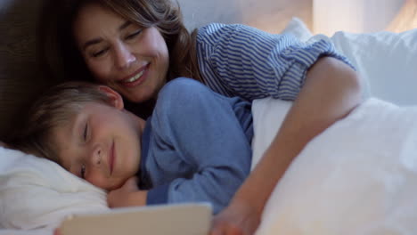Close-up-view-of-little-cute-boy-on-the-bed-while-his-pretty-mother-showing-him-a-tale-cartoon-on-the-tablet