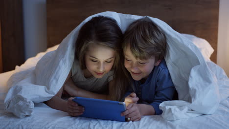 Cute-little-sister-and-brother-lying-under-the-blanket-and-watching-something-on-the-tablet-at-night