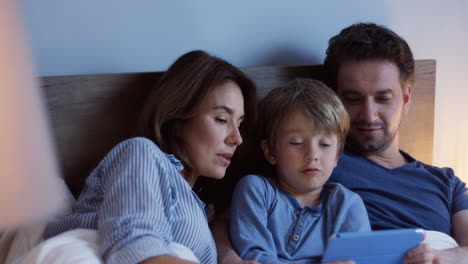 Close-up-view-of-caucasian-parents-sitting-on-the-bed-with-their-little-cute-son,-watching-something-on-the-tablet-and-talking-at-night