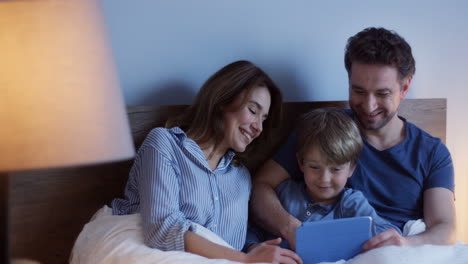 Caucasian-parents-sitting-on-the-bed-with-their-little-cute-son,-watching-something-on-the-tablet-and-talking-at-night