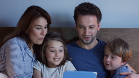 Close-up-view-of-caucasian-parents-sitting-on-the-bed-with-their-little-cute-son-and-daughter,-watching-something-on-the-tablet-and-talking-at-night
