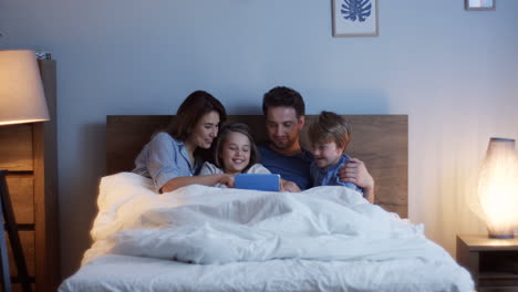 Caucasian-parents-sitting-on-the-bed-with-their-little-cute-son-and-daughter,-watching-something-on-the-tablet-and-talking-at-night