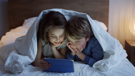 Cute-small-boy-and-girl-lying-under-the-blanket-and-watching-video-on-the-tablet-at-night