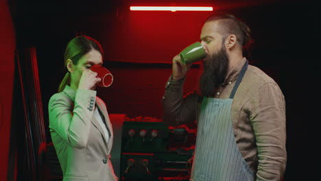 Caucasian-businesswoman-standing-in-metal-workshop-with-man-metal-industry-worker-with-beard-and-in-apron-talking-and-drinking-tea-or-coffee