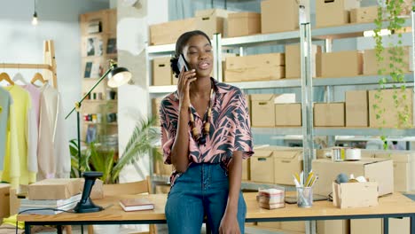 Mujer-Diseñadora-Afroamericana-Hablando-Por-Teléfono-Y-Apoyándose-En-La-Mesa-En-Una-Tienda-De-Ropa-De-Moda