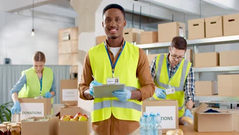 Front-view-of-African-American-male-volunteer-checking-donation-list-and-smiling-to-the-camera-in-warehouse-house