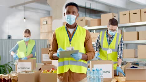 Front-view-of-African-American-male-volunteer-in-facial-mask-checking-donation-list-and-smiling-to-the-camera-in-warehouse-house