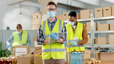 Front-view-of-caucasian-male-volunteer-in-facial-mask-checking-donation-list-and-smiling-to-the-camera-in-charity-warehouse