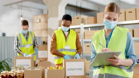 Front-view-of-caucasian-female-volunteer-in-facial-mask-checking-donation-list-and-smiling-to-the-camera-in-warehouse-house