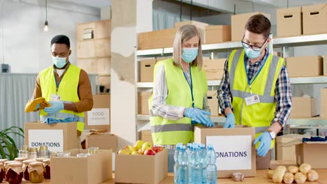Multiethnic-group-of-senior-and-young-volunteers-in-facial-masks-packing-food-and-clothes-in-donation-boxes-in-charity-warehouse