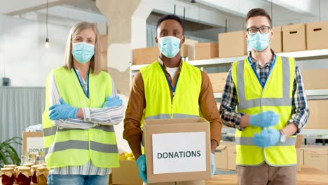 Multiethnic-group-of-senior-and-young-charity-workers-in-facial-masks-holding-donation-box-and-looking-at-camera-in-charity-warehouse