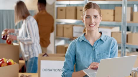 Close-up-view-of-caucasian-young-female-volunteer-typing-on-laptop-and-smiling-to-the-camera-while-her-coworkers-packing-donation-boxes-in-charity-warehouse