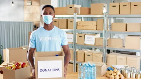 Afroamerikanischer-Männlicher-Freiwilliger-Mit-Gesichtsmaske,-Spendenbox-Und-Lächeln-In-Die-Kamera-Im-Wohltätigkeitslager