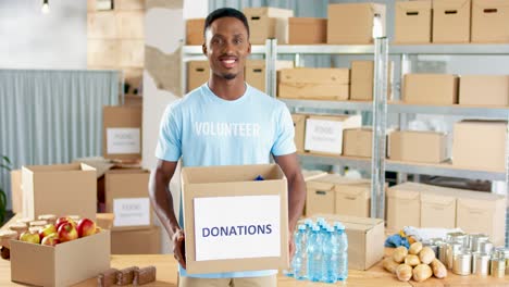Voluntario-Afroamericano-Sosteniendo-Una-Caja-De-Donación-Y-Sonriendo-A-La-Cámara-En-Un-Almacén-De-Caridad