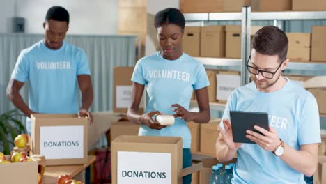 Multiethnische-Gruppe-Von-Freiwilligen,-Die-Kartons-Mit-Lebensmitteln-Und-Kleidung-Packen-Und-In-Einem-Lager-Für-Wohltätigkeiten-Auf-Einem-Tablet-Tippen